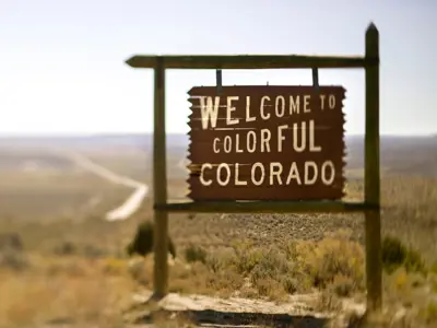 A weathered wooden sign reads "Welcome to Colorful Colorado" against a vast, sunlit landscape, evoking a sense of adventure and exploration.