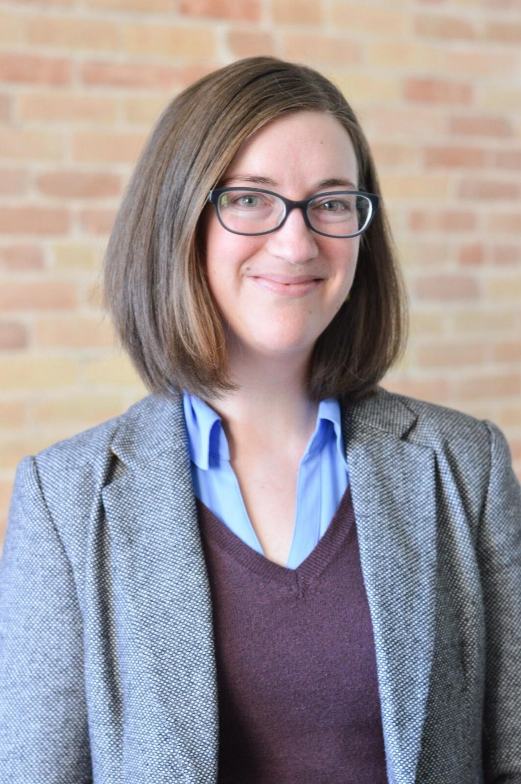 Headshot of a woman wearing glasses and a grey suit against a brick wall.
