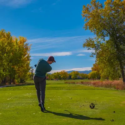 A man swings a golf club, with trees and a blue sky in the background.