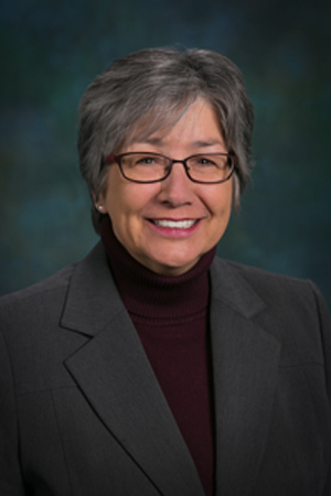 Headshot of a woman wearing glasses and a dark suit against a grey background.